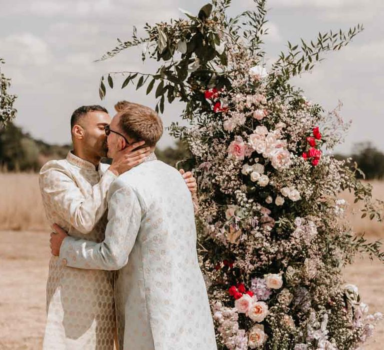 Groom in mint green Indian Sherwani with white garden rose and foliage boutonniere and grooms sunglasses doing first kiss as husbands with groom in cream and gold Indian sherwani and white garden rose boutonniere 