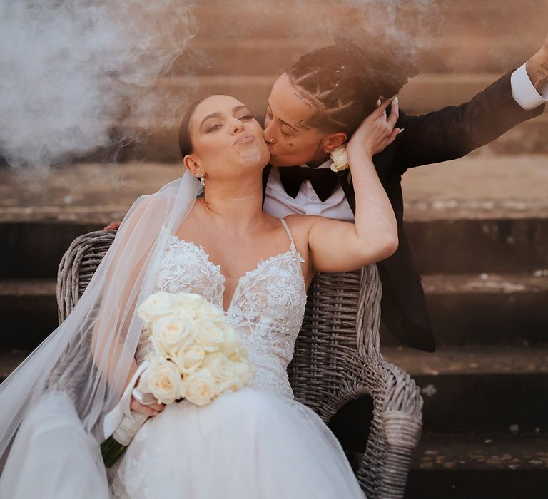 Bride in black snakeskin print tuxedo with black bowtie kissing bride on the cheek in lace sleeveless v neck Enzoani wedding dress with tulle finish during wedding smoke bomb photoshoot 