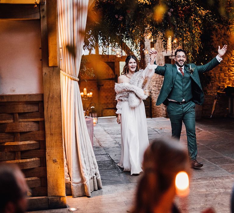 Bride walking into the wedding reception at Cripps Barn with faux fur shawl with the groom in autumnal three piece suit 