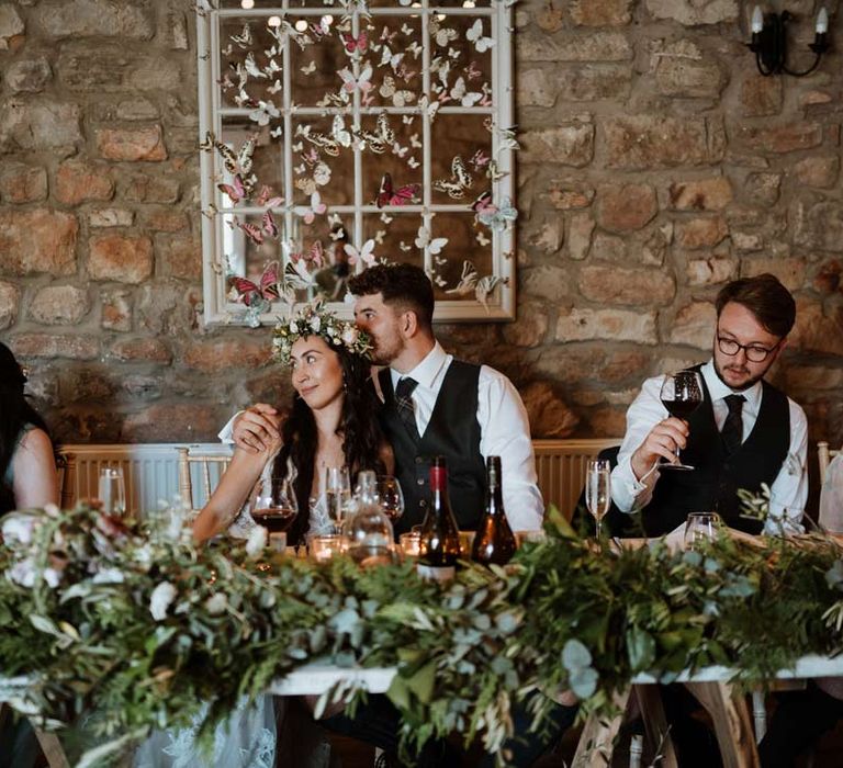 Bride in white garden rose bridal flower crown sitting with groom in dark grey waistcoat and tartan tie at The Barn at Harburn wedding venue with foliage centrepieces and 3d paper butterfly decorations