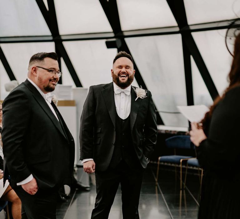 Grooms wearing three piece black tuxedos with off white bow ties and white garden rose, lavender twig and dried foliage boutonnieres at the alter with wedding celebrant at The Gherkin London 