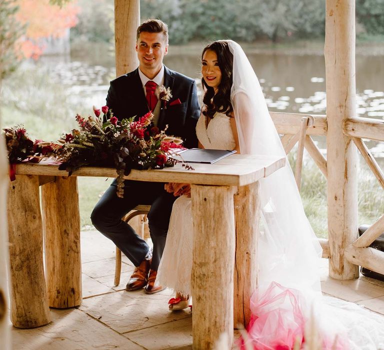 Bride in sleeveless lace wedding dress and church length red ombre veil sitting with groom in deep blue grooms blazer, grey waistcoat, red tie, red pocket square and mixed dried flower boutonniere 