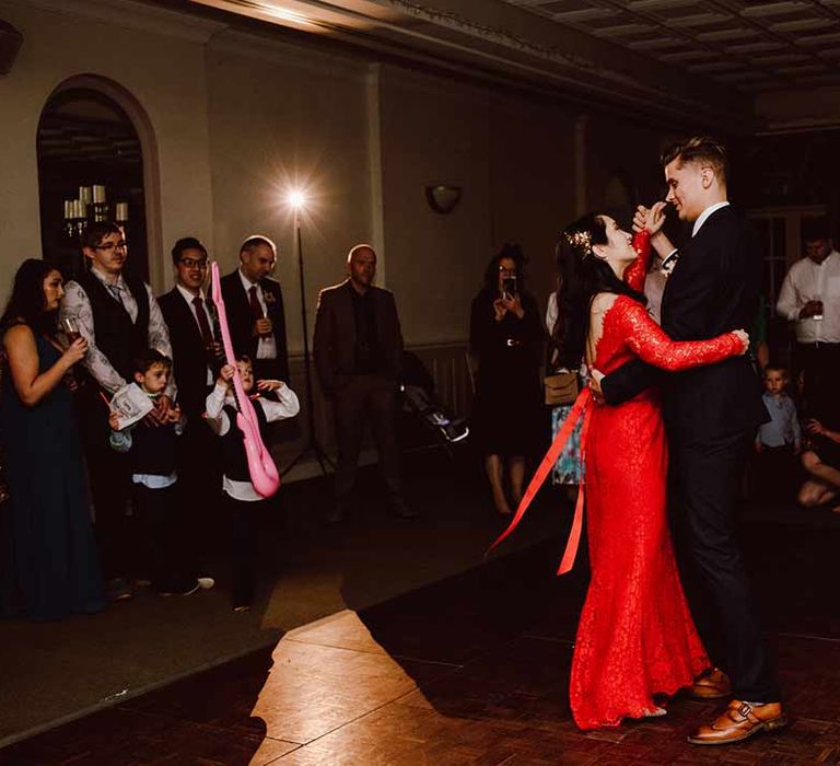 Groom wearing deep blue suit doing wedding first dance with bride in red lace wedding dress with long sleeves, red and gold hair accessories and golden closed toe wedding heels