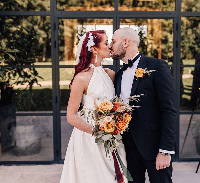 Groom in black tux kissing the bride in a halter neck wedding dress from Halfpenny London 