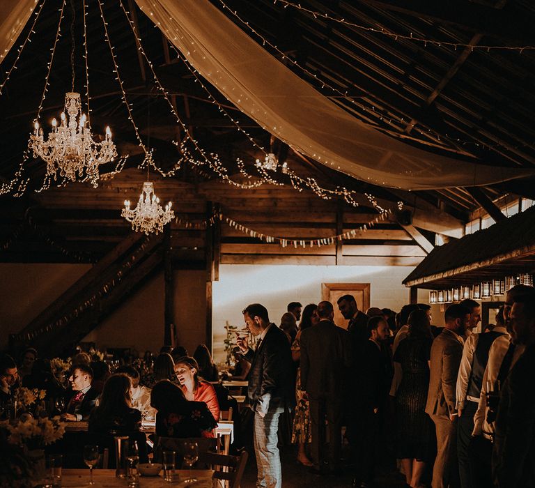 The wedding guests stand chatting, eating, and drinking with white drapery and fairy lights for cosy rustic barn wedding 