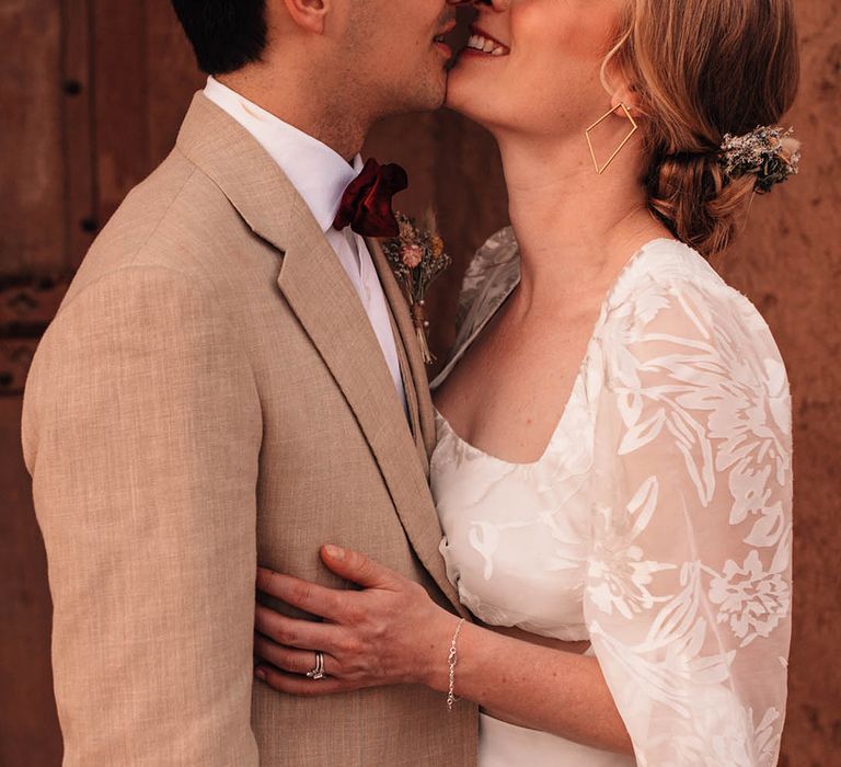 Bride in lace long sleeve crop top with the groom in cream suit leaning in for a kiss 