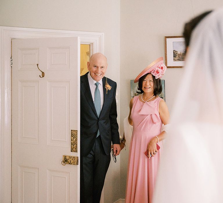 Mother of the bride in full pink outfit with the father of the bride looking at the bride in her classic wedding dress 