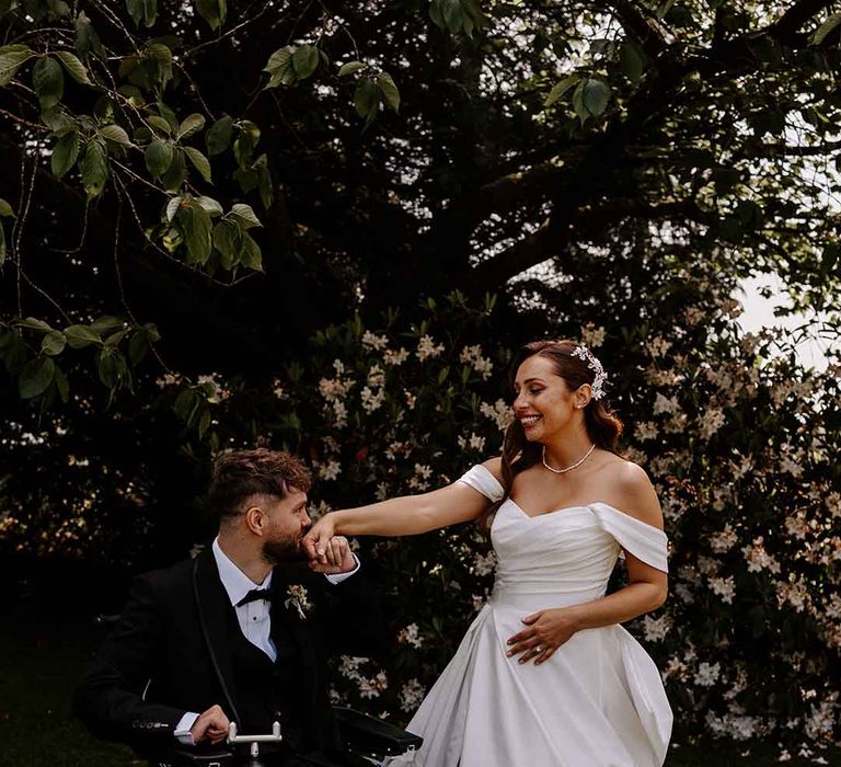 groom wearing a tuxedo in a wheelchair kissing his bride's hand in an off the shoulder wedding dress