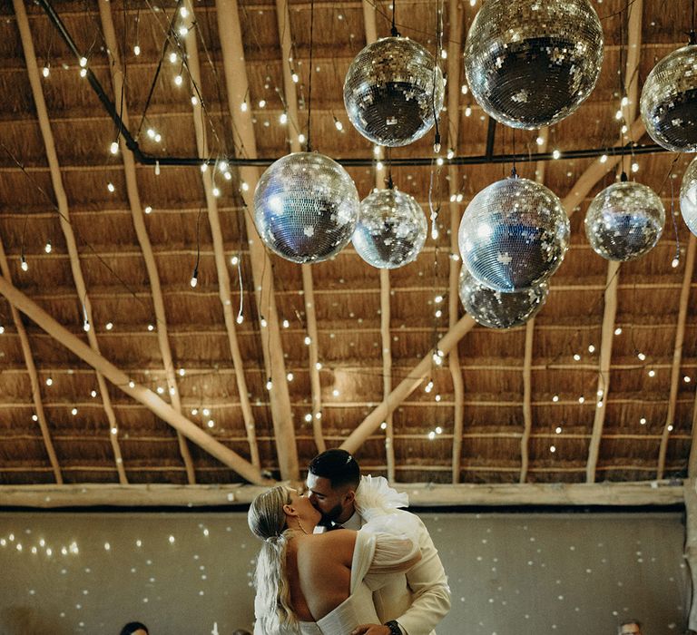 bride in a mermaid wedding dress kissing her groom in a white tuxedo jacket on the dance floor under a canopy of disco balls 