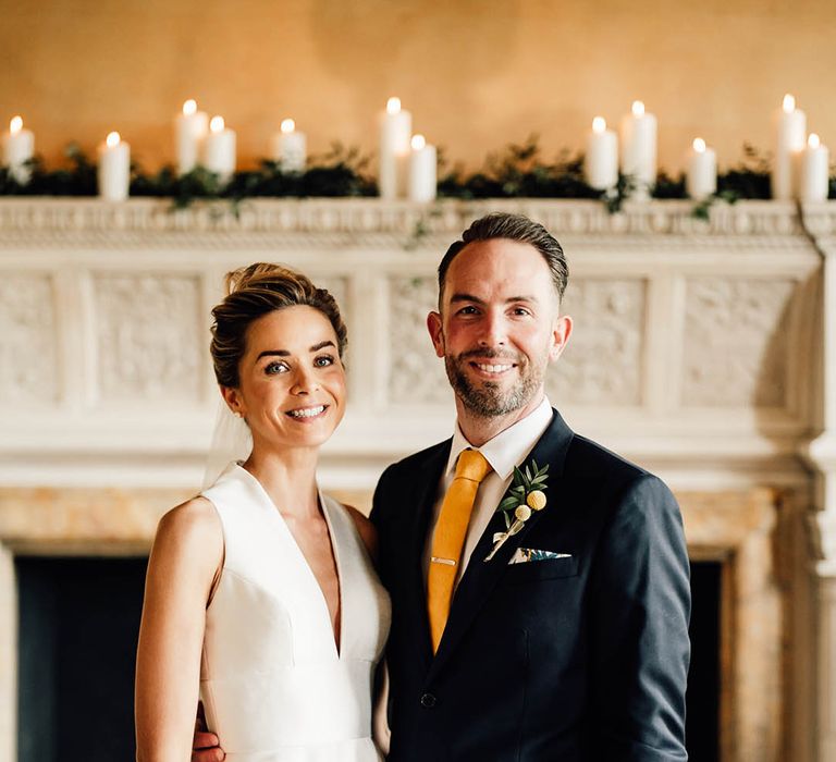 Bride in a plunging neckline Sheila Harding sleeveless wedding dress with the groom in a navy suit with yellow tie and buttonhole with tie clip 