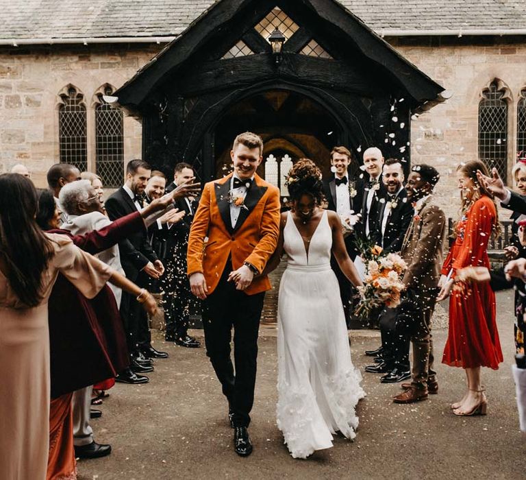 Bride in v-neck sleeveless wedding dress with 3D floral applique train holding blush and orange garden roses, poppies and carnations, dried flowers and eucalyptus wedding bouquet walking with groom in crushed burnt orange velvet grooms blazer, black bowtie and black suit trousers doing colourful confetti exit