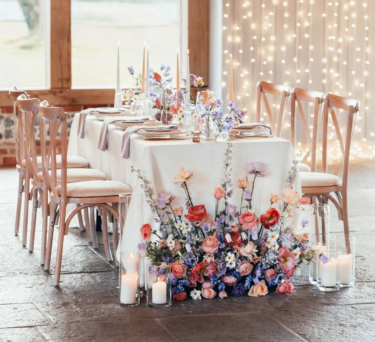 Peach and lilac wedding tablescape with spring flower centrepieces, lilac napkins, tinted glassware and gold cutlery and crockery on white tablecloth at Rackleys Barn with fairy lights wall behind