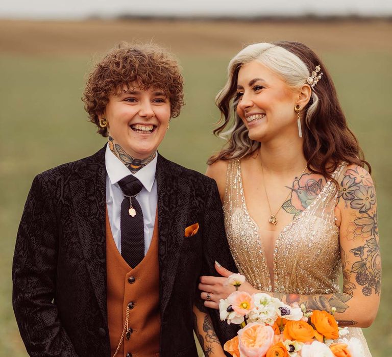 Bride in dark velvet bridal jacket with rust waistcoat and pocket square, black suit trousers and gold pocket watch standing with bride in sleeveless deep v-neck sparkly gold wedding dress holding orange garden rose, white poppy, light pink carnation and foliage bridal bouquet wrapped in sparkly gold ribbon at Kingscote Barn Tetbury wedding