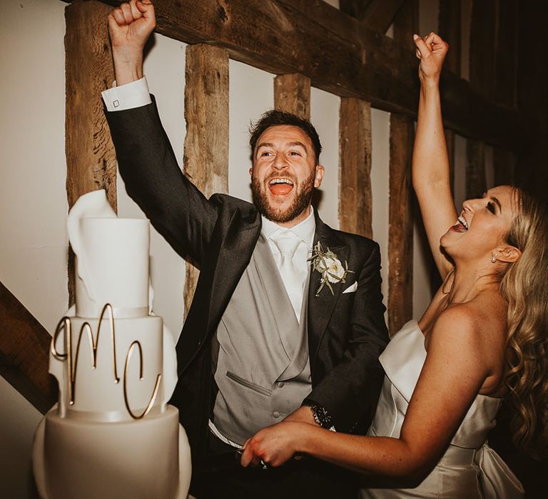 The bride and groom celebrate as they go into cut the wedding three tier white iced wedding cake with gold wire initial letters 