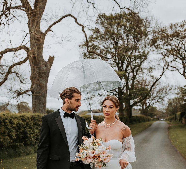 Groom in black tuxedo carrying clear umbrella with bride in corset style Made With Love wedding dress with neutral blush bouquet 