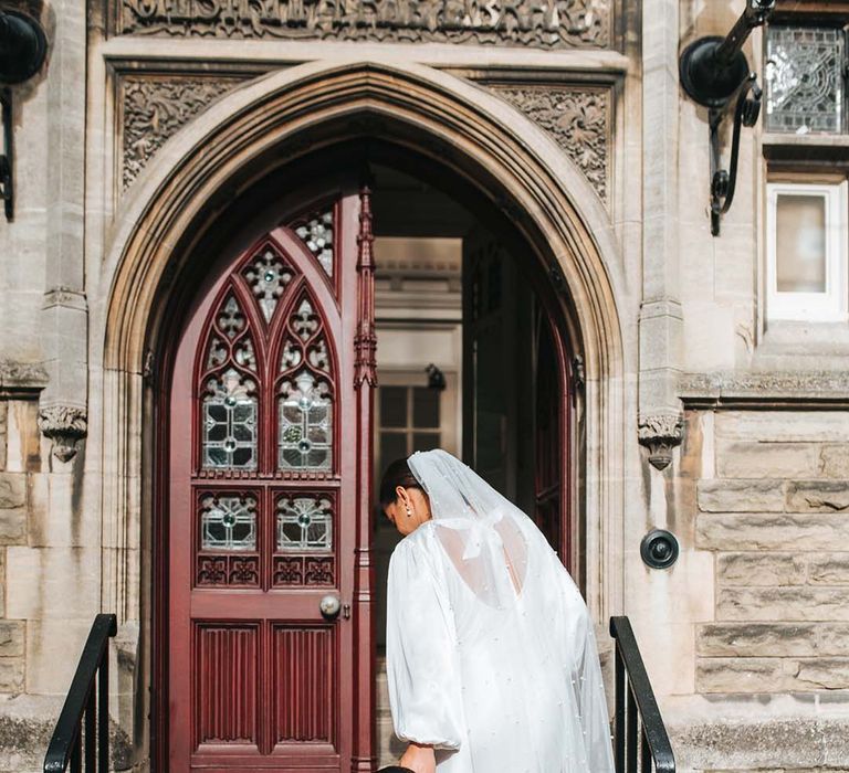 Bride in long sleeve satin wedding dress with pearl embellished veil walking up the stairs to the registry office with her son