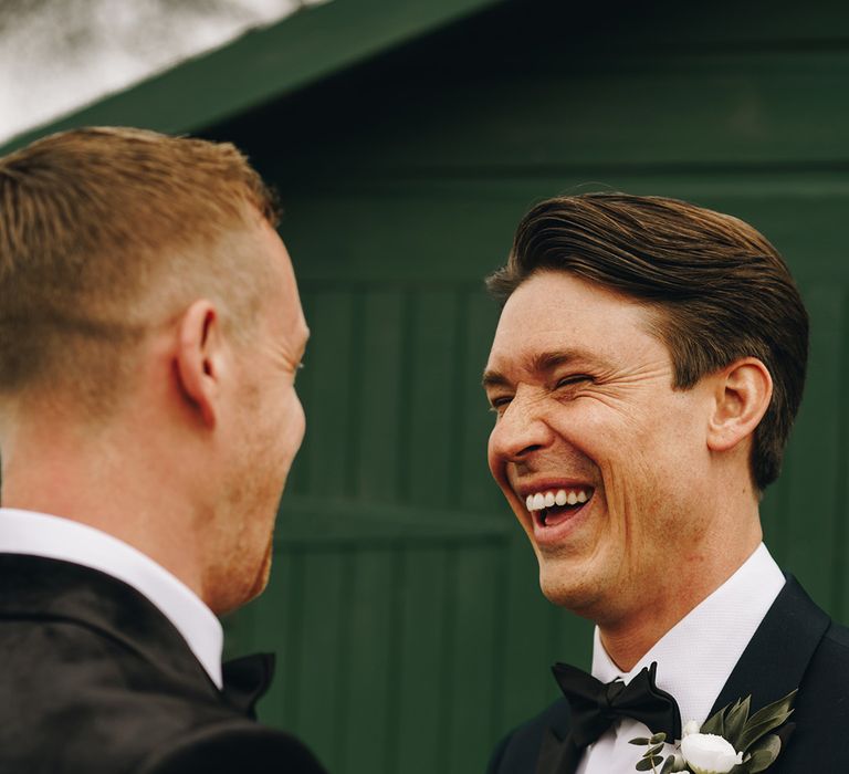 Groomsman puts on white flower buttonhole onto the groom's black tuxedo 