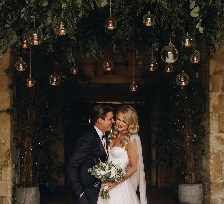 Bride with wavy blonde hair wearing sparkly corset bodice wedding dress with groom under hanging installation at Stone Barn