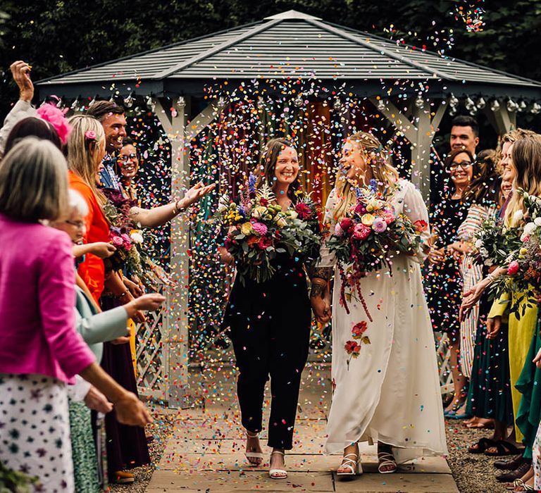 Outdoor wedding ceremony for two brides who walk back down the aisle as colourful confetti is thrown over them 