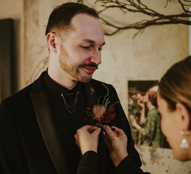 The groom in a velvet suit jacket gets help fixing on his dried flower buttonhole accessory 