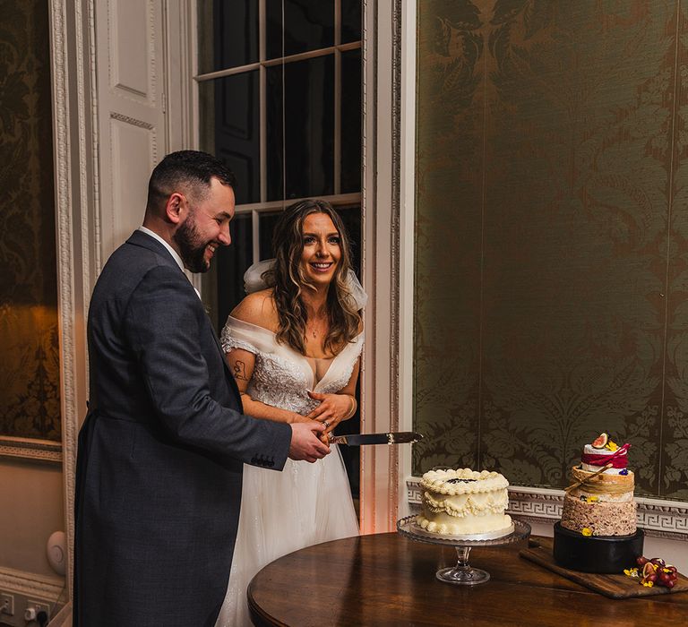 Groom in navy morning suit cutting the retro wedding cake with the bride 