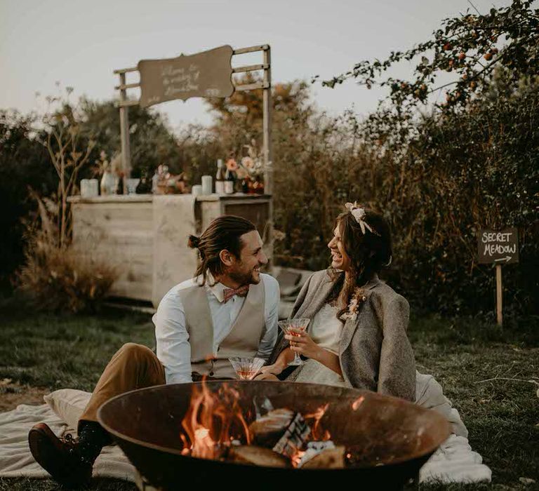 The bride and groom sit in front of the fire pit at their outdoor wedding