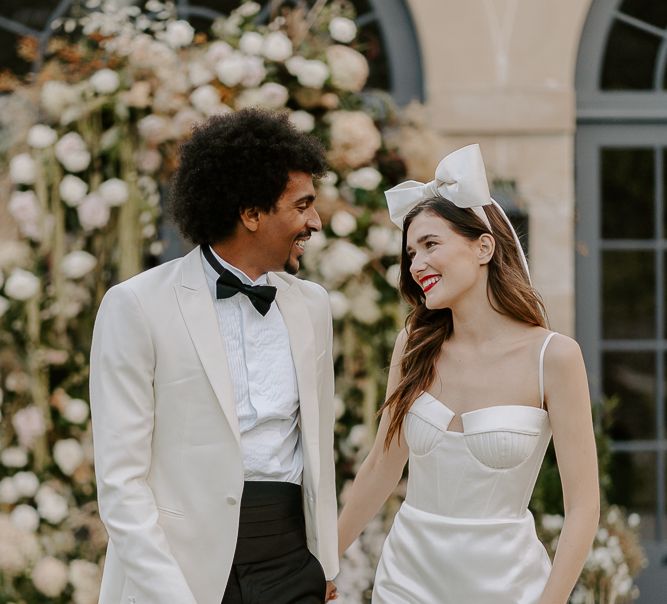 Black groom with afro hair and white tuxedo jacket holding hands with his bride with long brown hair, red lipstick and bow headdress walking down the aisle 