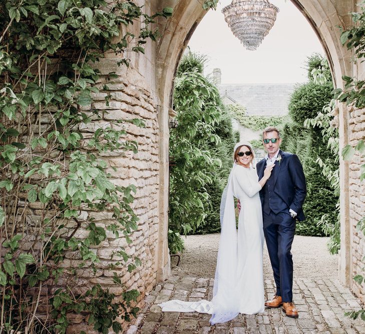 Bride in Elbeth Gillis gown stands in arms of groom in navy Cad & the Dandy suit and sunglasses under chandelier hanging in archway outside at Euridge Manor