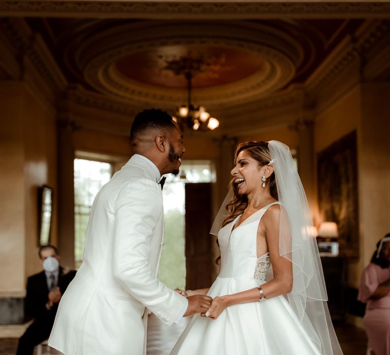 Groom in a white tuxedo jacket smiling at the altar with his bride in a Justin Alexander princess gown 