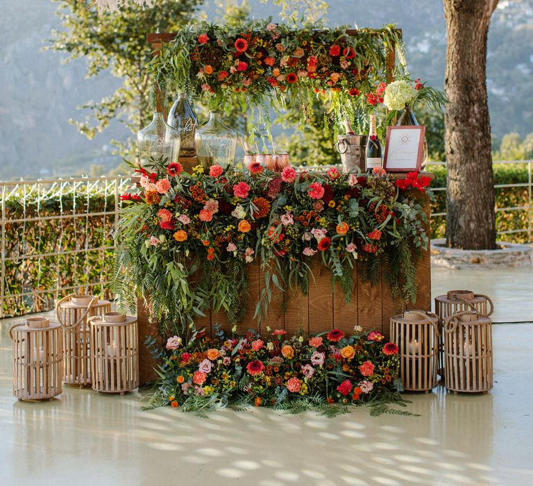 An outdoor cocktail bar covered in orange, red and pink flowers to match the bride's bouquet, with lanterns on either side