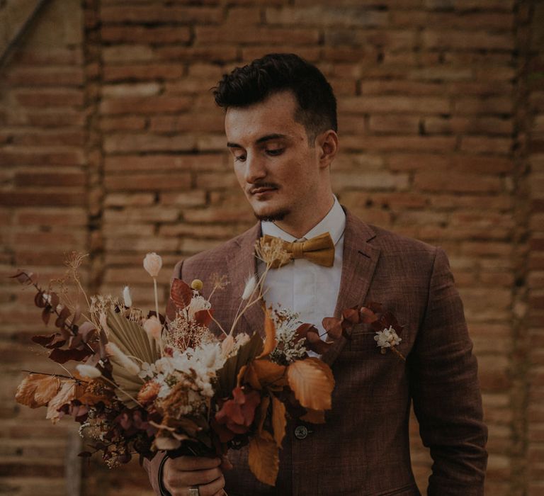 Groom in a brown wedding suit and orange bow tie holding an autumn leaf and flower wedding bouquet 