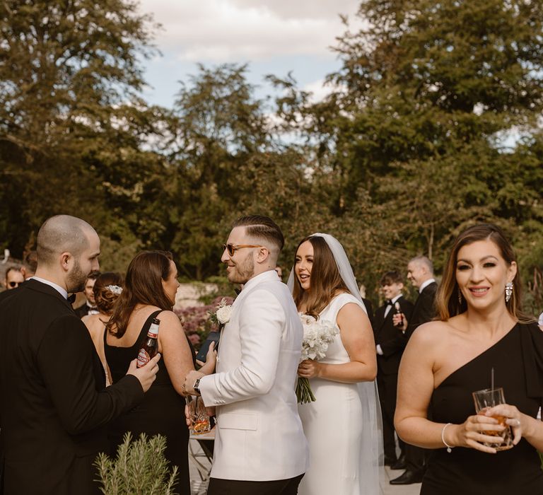 Groom in white tuxedo jacket and sunglasses and bride in Made With Love wedding dress greet guests outside the Fig House at Middleton Lodge