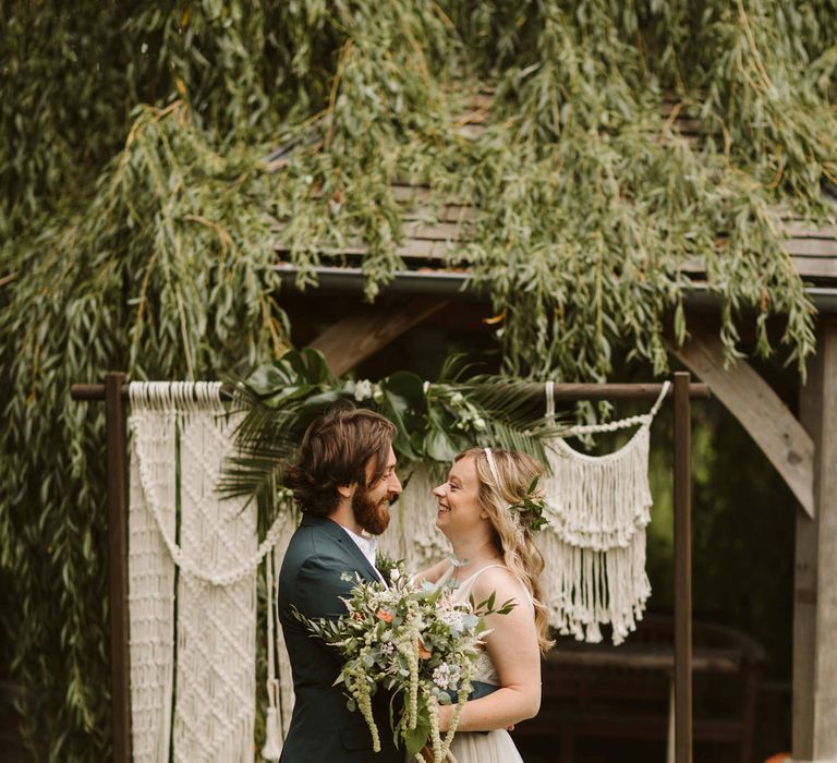 Bride & groom stand in front of macrame decor whilst bride holds bouquet tied with ribbon