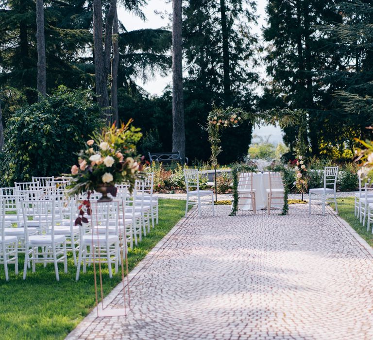 A beautiful outdoor wedding ceremony with floral plinths, white seating and a flower archway