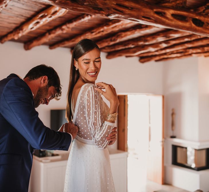 Groom in a navy suit helping his bride into an embroidered Marylise Bridal gown with long sleeves