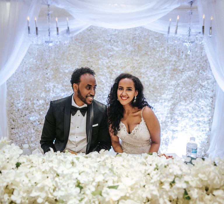 Bride and groom sitting at the top table with all white flower wall and top table arrangement 