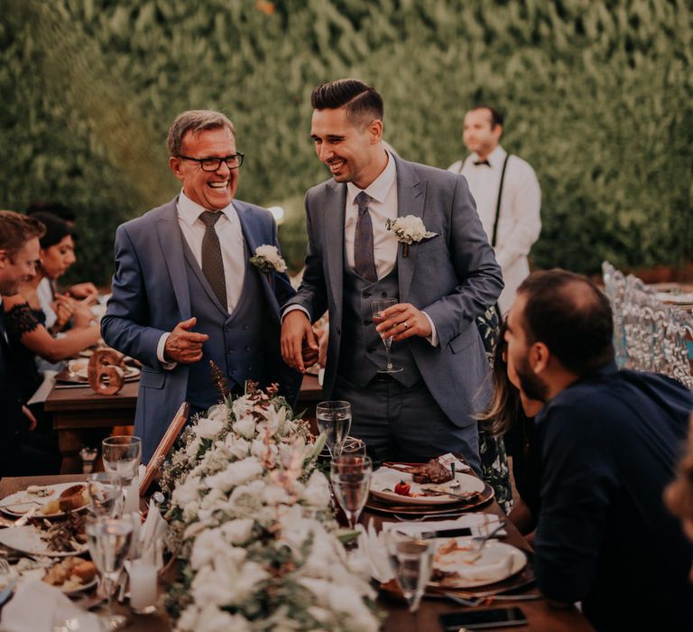 The groom and his father laughing at a table wearing matching blue three piece suits