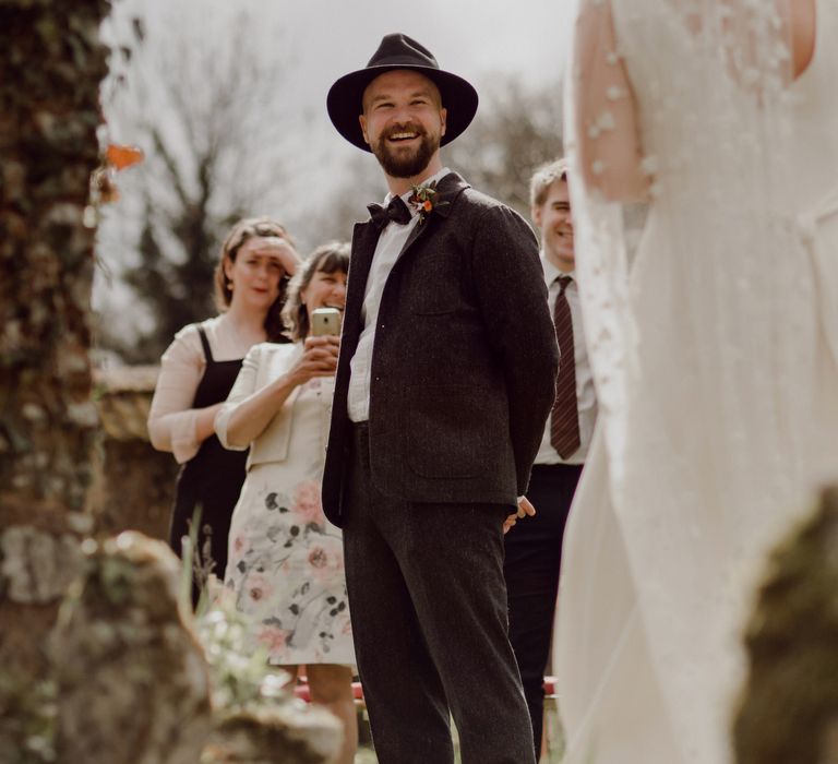 Groom in grey tweed suit and black fedora smiles at bride in Charlie brear wedding dress and chapel length daisy applique veil walking down the aisle at garden party wedding