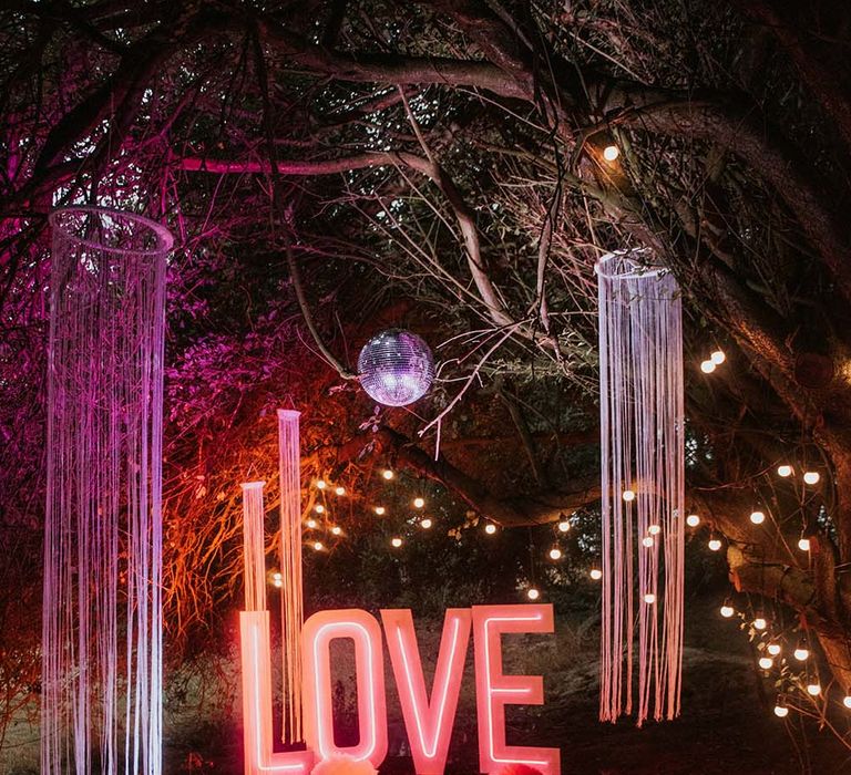 Pink neon sign spelling LOVE with hanging streamer lights and disco ball outside at festival themed wedding 