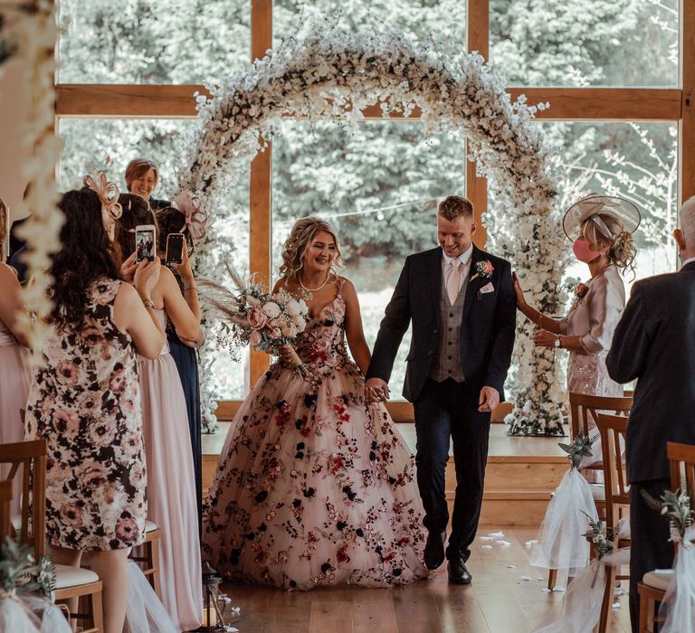 Bride & groom walk down the aisle after wedding ceremony with floral arch in the background