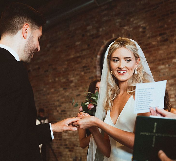 Beautiful bride with half up half down wedding hair wearing a veil putting on her grooms wedding band 