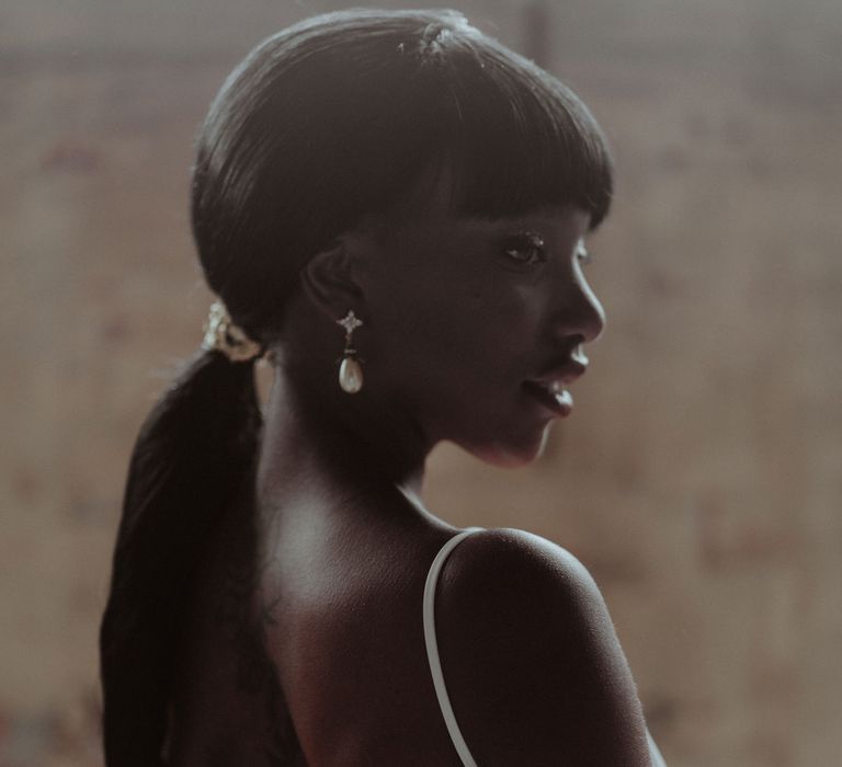 A Black bride stands with her back to the camera showing off a low back white wedding dress. Her long hair is back in a low ponytail. 