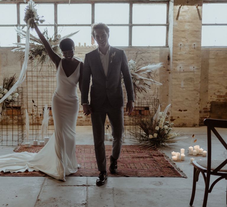 A bride and groom walk down the aisle in an industrial wedding venue. She raises her bouquet in the air.