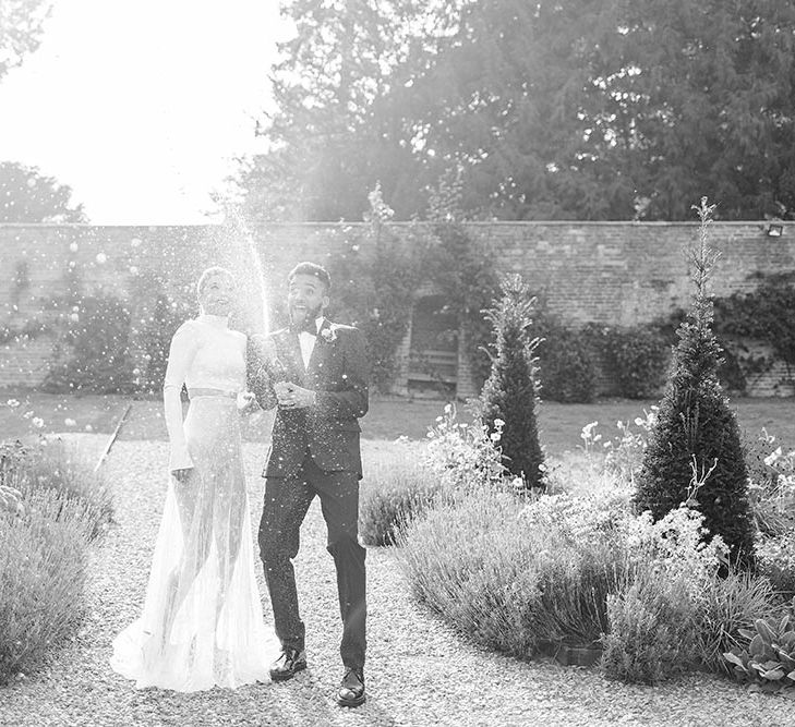 Bride in separates and groom in a tuxedo spraying champagne in the gardens at Garthmyl Hall