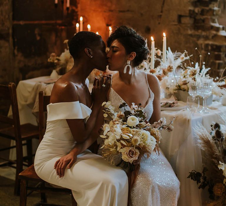 Two brides sitting at their reception table kissing and holding a dried and fresh flower neutral wedding bouquet 