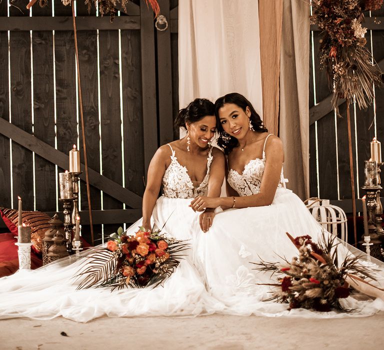 Two brides in tulle skirt dress with lace bodices sitting on the floor at The Barn at Drovers with dried and fresh flower installations and candle and lantern floor decor 