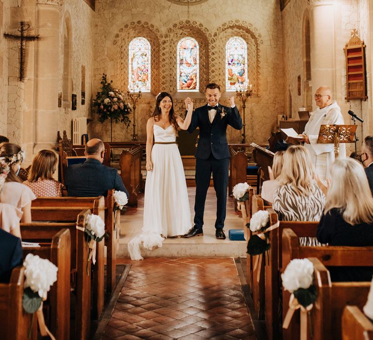 Bride in white cami wedding dress with train and veil celebrates holding the hand of groom in blue suit and bow tie at the altar after wedding ceremony