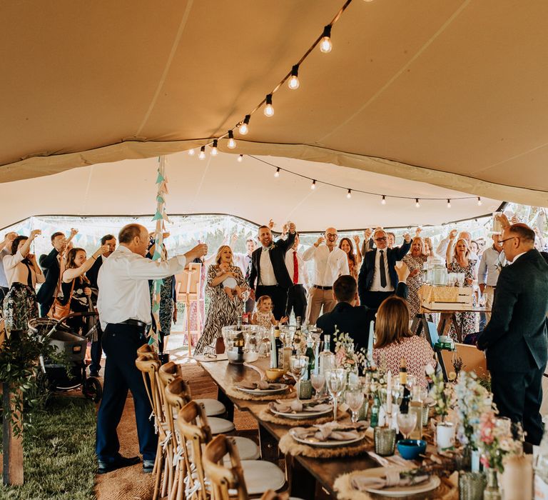 Interior of tipi with string fairy lights at garden wedding reception with rustic wedding breakfast tables with guests raising a glass