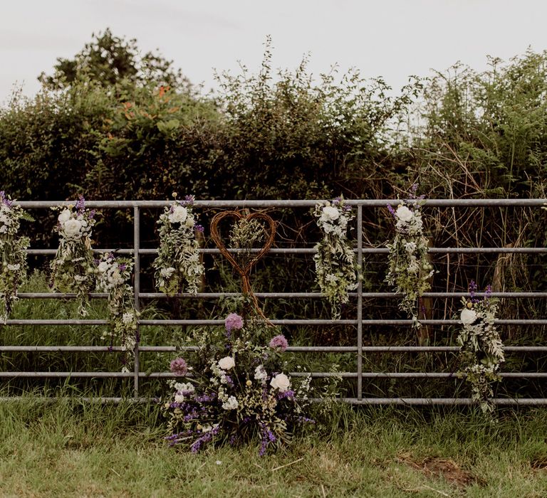 Metal farm gate with white and purple florals attached and wooden heart at home farm wedding