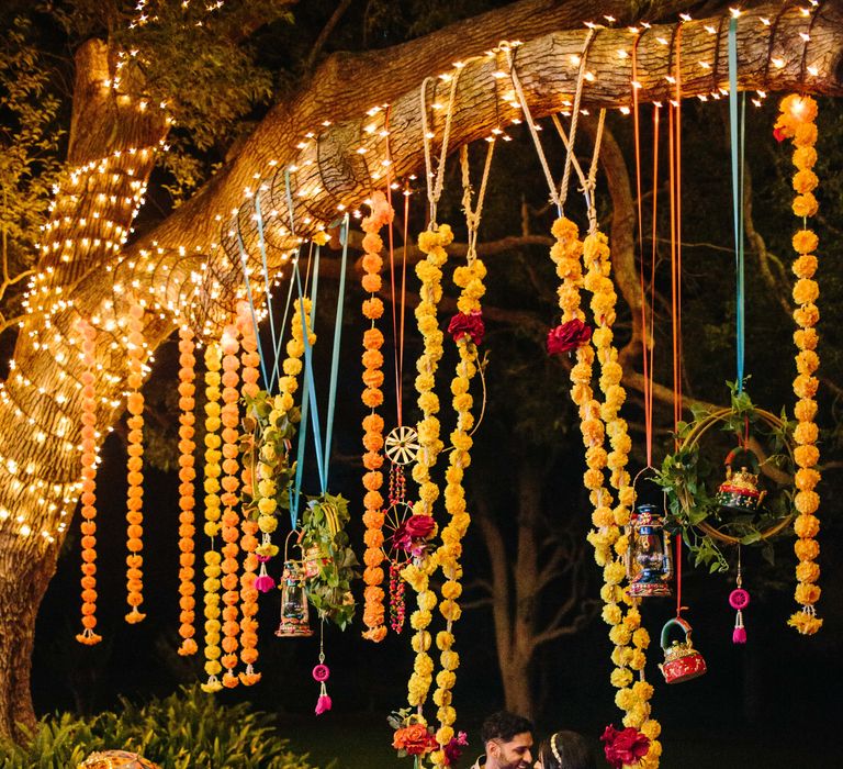 Colourful decor hangs from trees as bride & groom sit on swing and look lovingly at one another outdoors